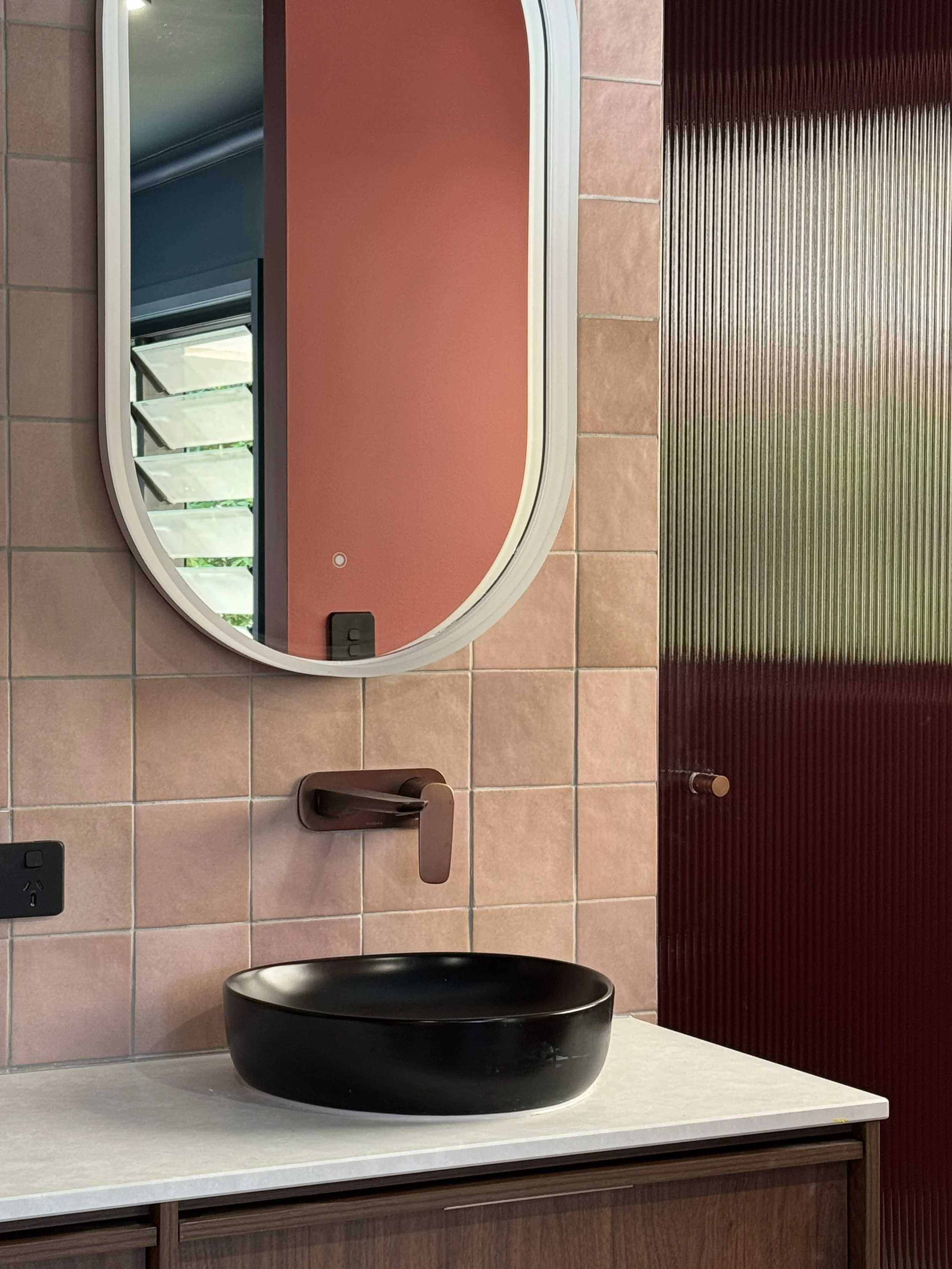 A modern bathroom sink area with a black vessel sink on a wooden cabinet, a bronze faucet, a large curved mirror, pink tiled wall, and a textured dark red wall with a towel hook.