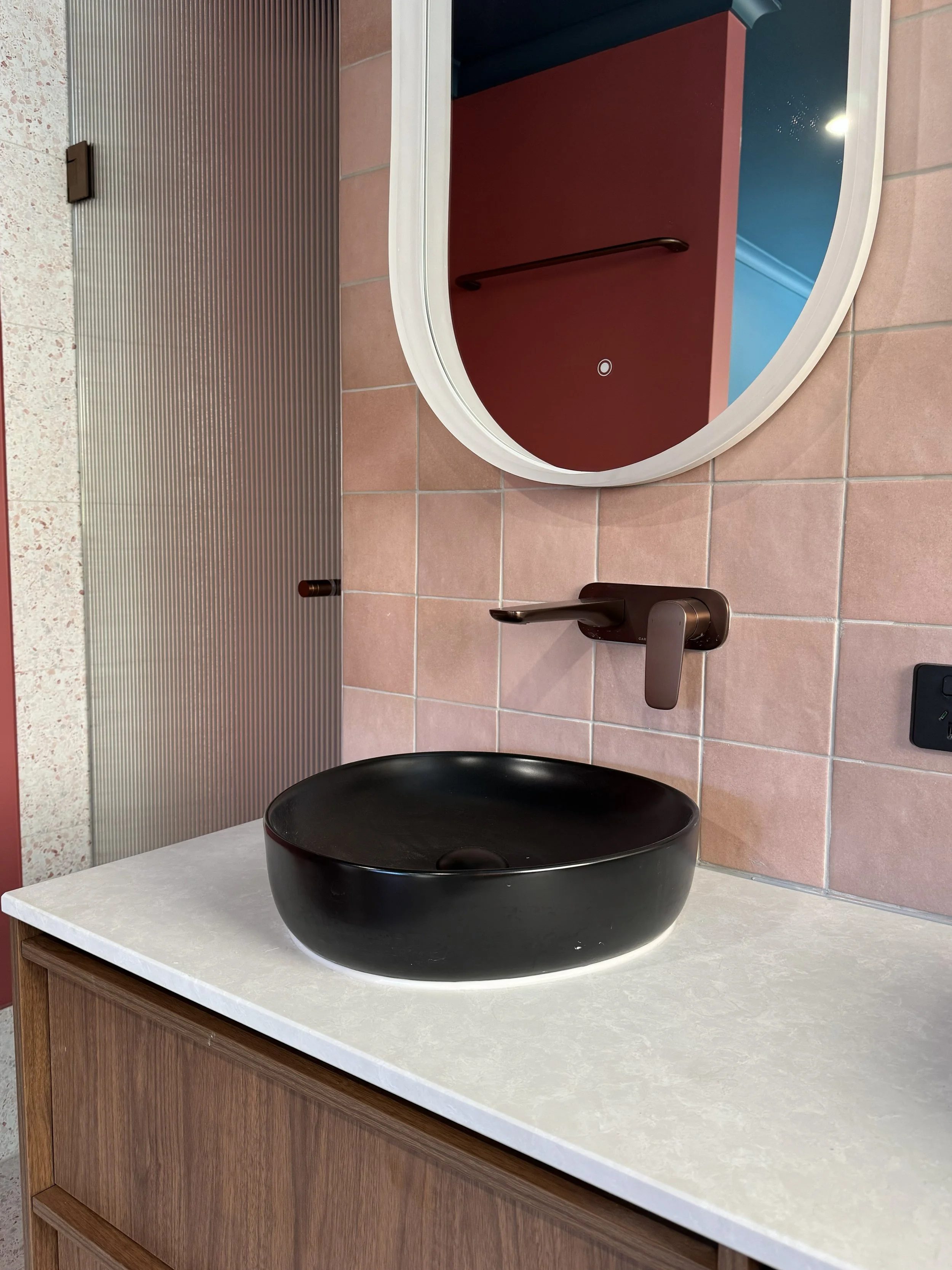 Black vessel sink on a white countertop with a bronze everyday faucet above it, pink tiled wall behind, a round mirror reflecting a red wall and blue ceiling, and metallic wall panel on the side.
