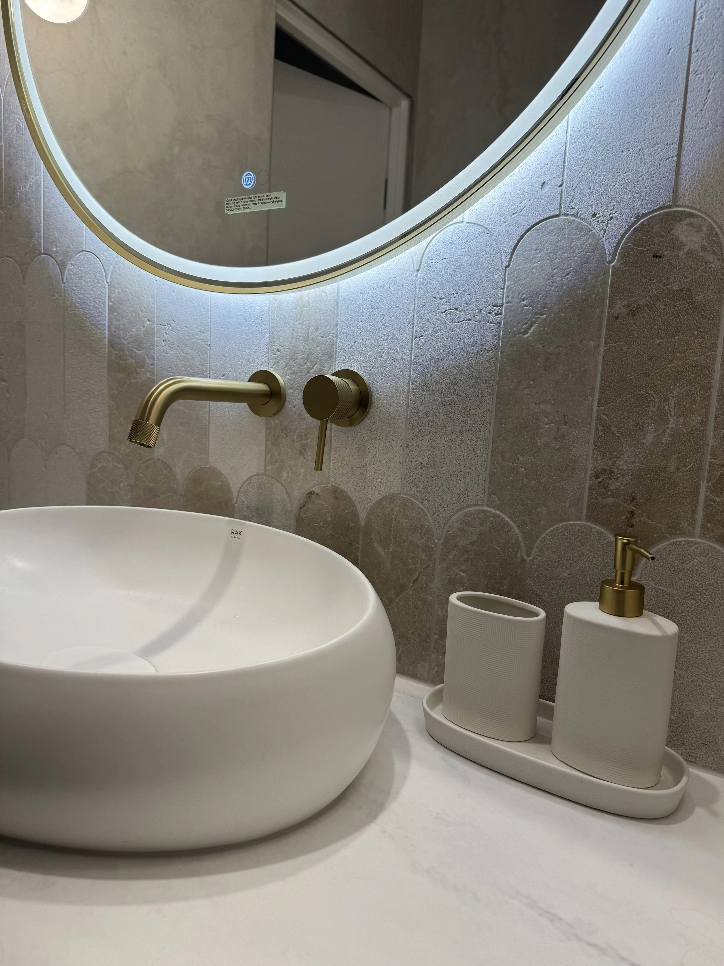 Close-up of a modern bathroom sink area with a circular mirror, wall-mounted gold faucet, soap dispenser, and toothbrush holder on a white countertop. The mirror has LED lighting around its edge and the wall behind features textured, decorative tiles