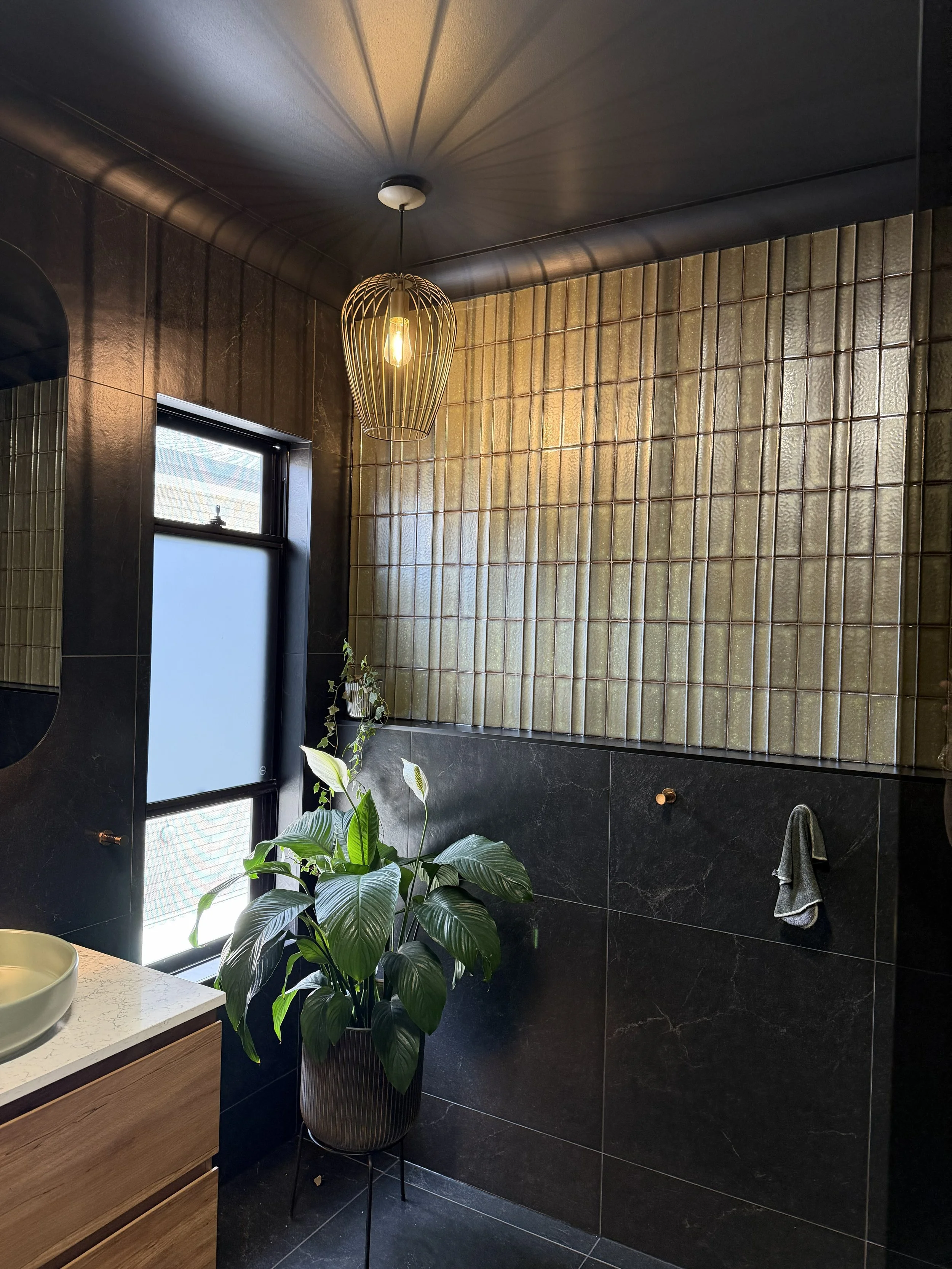 A modern bathroom with a black tiled wall, a large window with frosted glass, a potted green plant, and a gold pendant light fixture hanging from the ceiling.