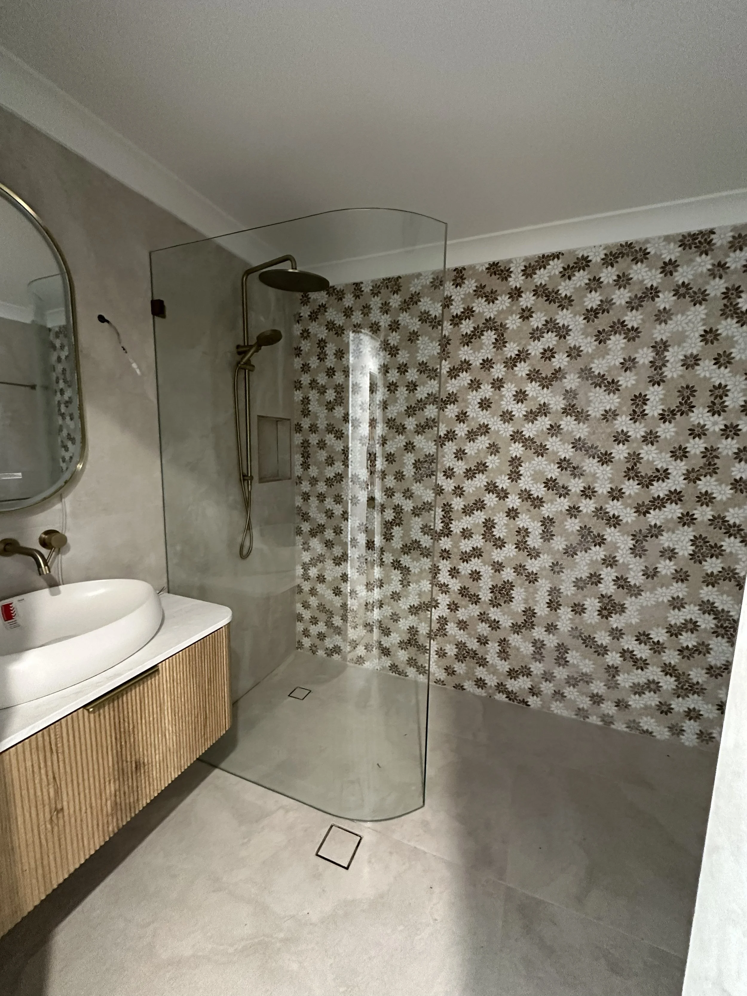 Modern bathroom with a walk-in shower, floral patterned wall, white oval sink, mirror, and wooden vanity.