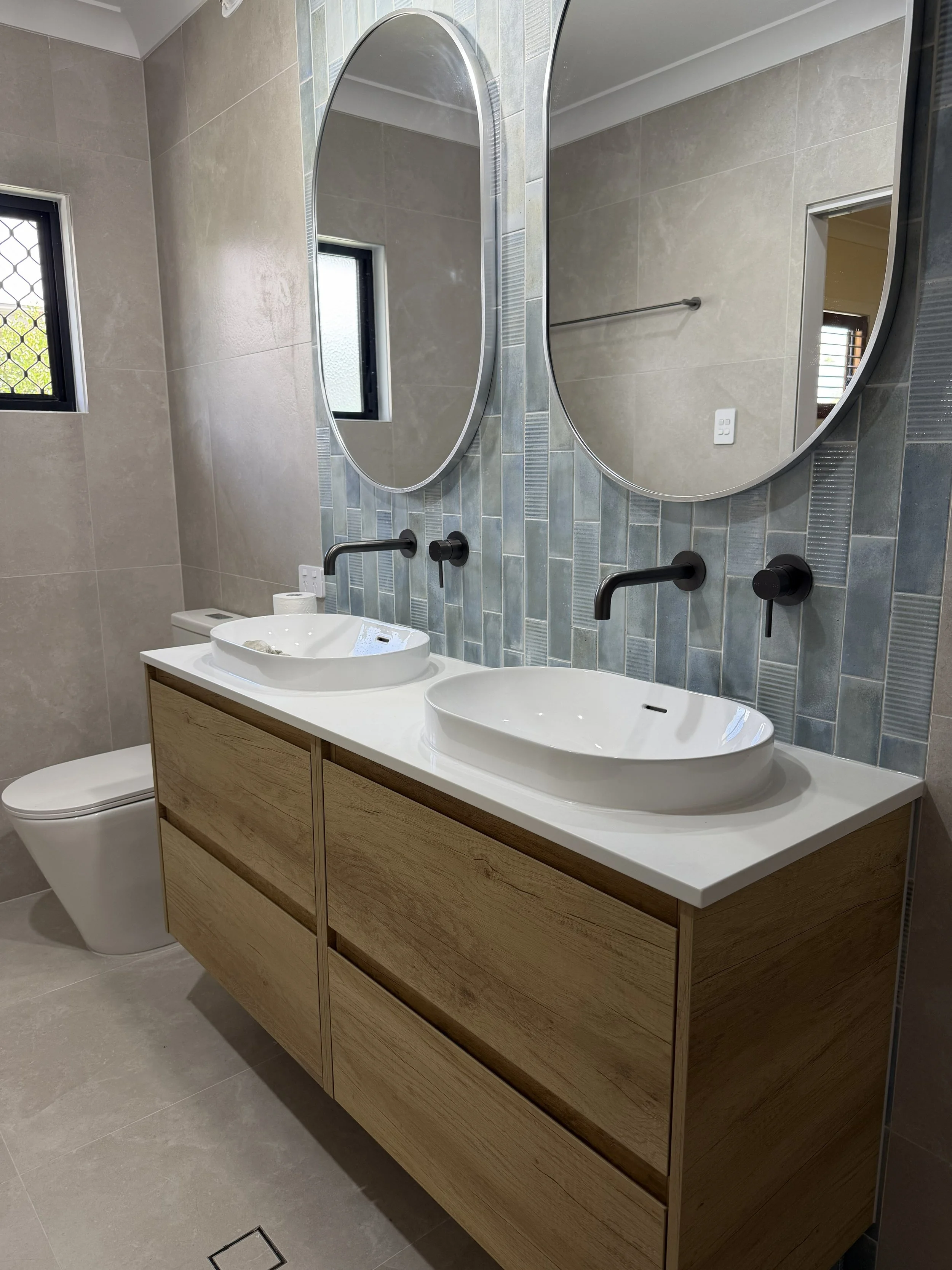 Modern bathroom with dual white vessel sinks, black wall-mounted faucets, a wooden vanity, two oval mirrors, a small window, and beige tiled walls and floor.