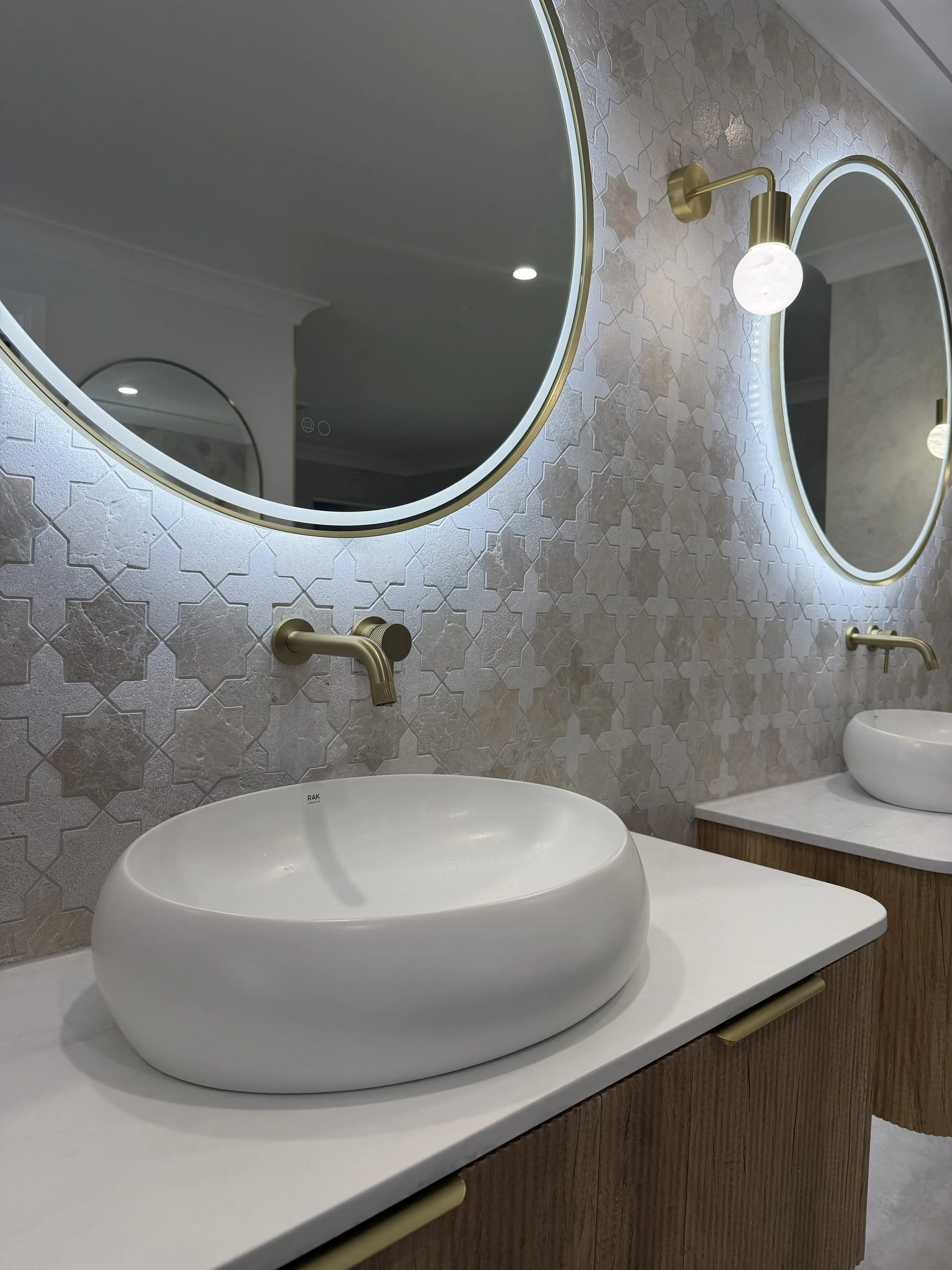 Modern bathroom with round white vessel sinks, brass faucets, large backlit oval mirrors, and beige patterned wall tiles.