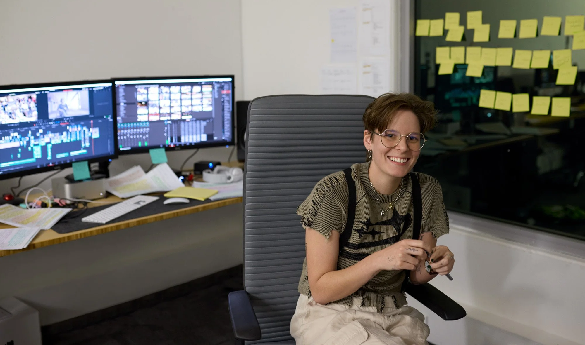 A young woman with short brown hair, glasses, and earrings smiling while sitting in an office chair. She is wearing a tan and black patterned shirt and beige pants. Behind her is a desk with dual computer monitors displaying video editing software, scattered papers, sticky notes, and office supplies. To the right, yellow sticky notes are arranged on a glass wall.