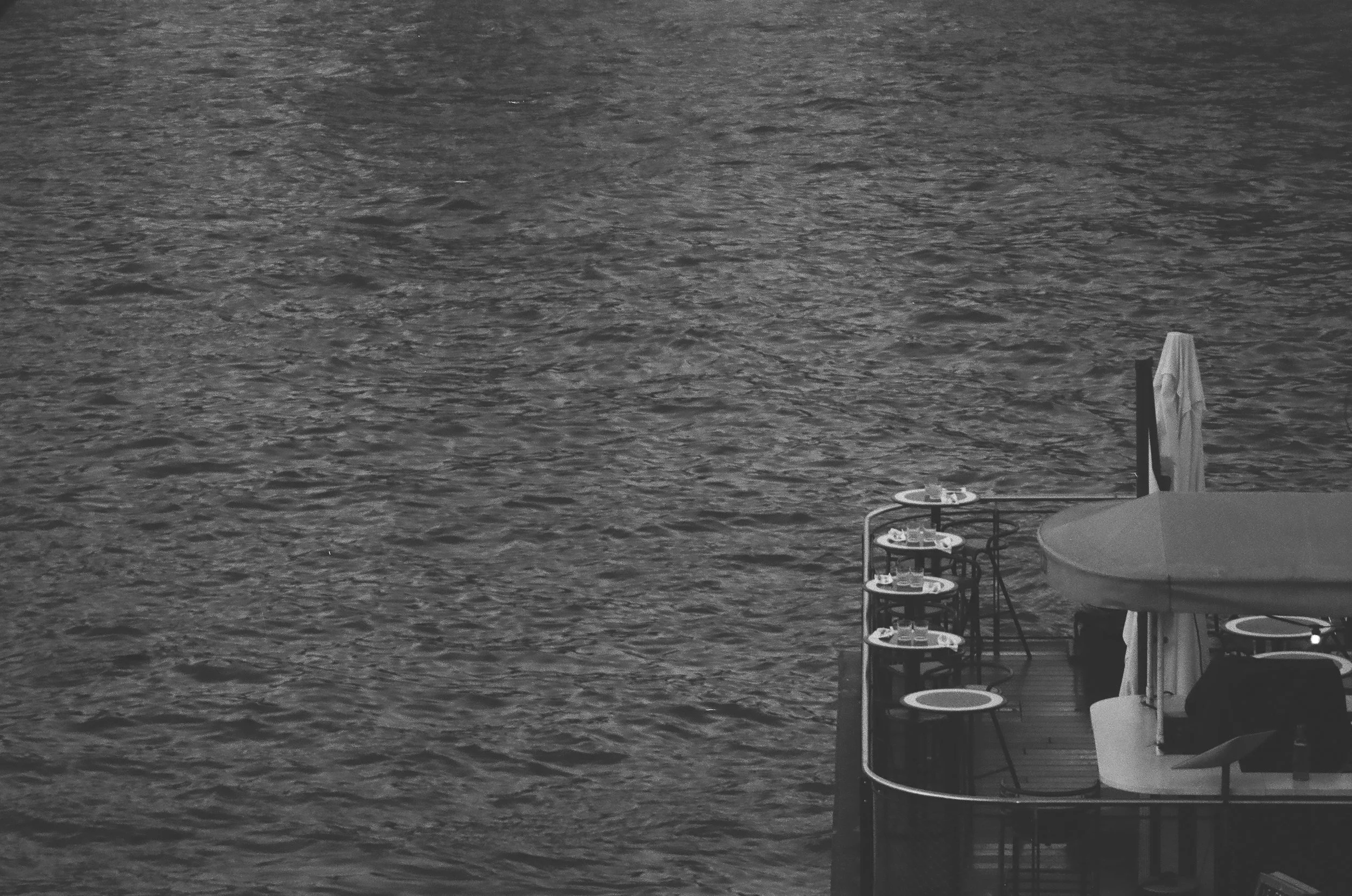 A black and white photograph of a body of water with a patio area on the right side, featuring tables, chairs, and a umbrella.