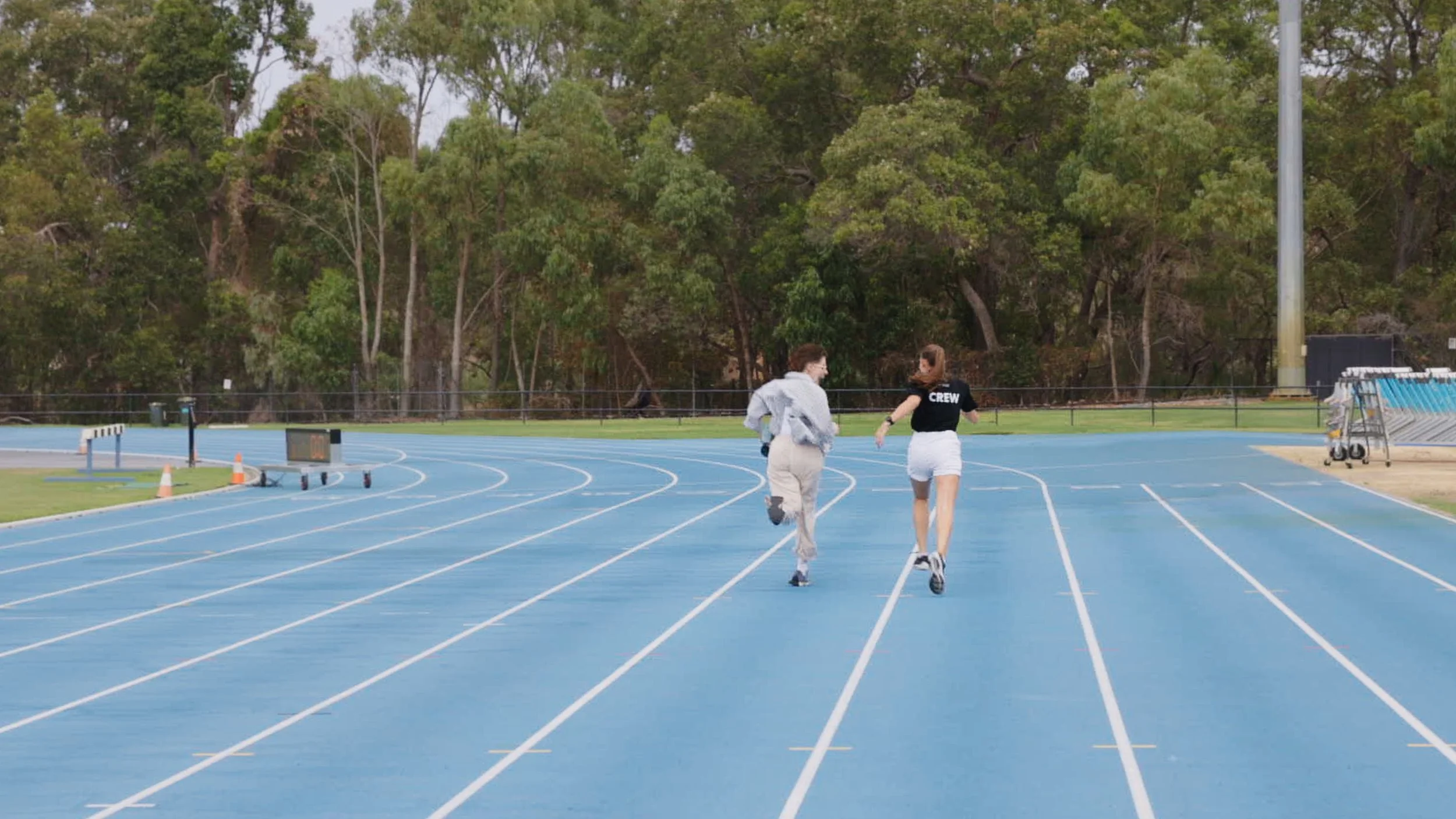 Two people running on a blue outdoor track with a lush green forest in the background.