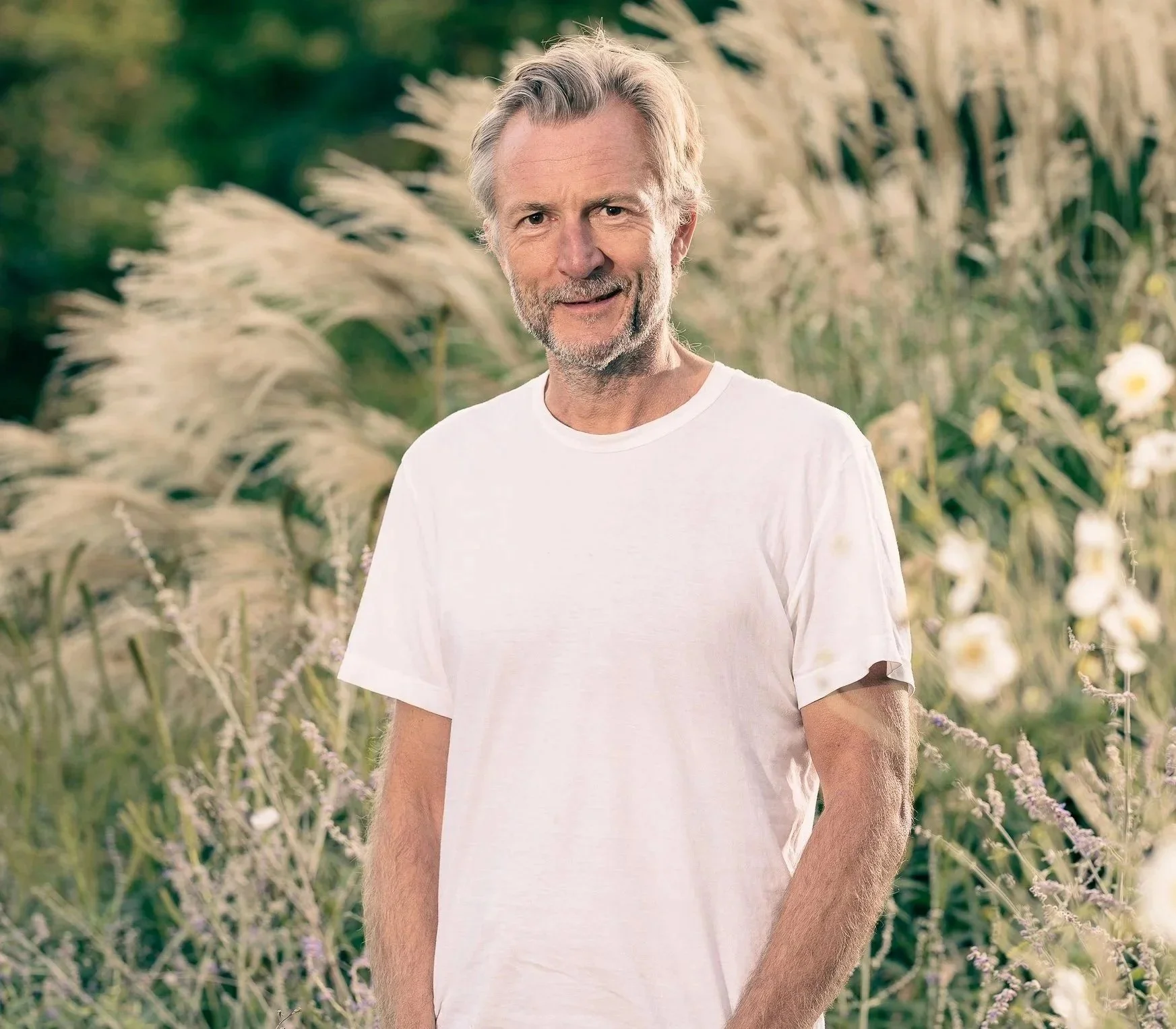 A smiling older man with gray hair and a beard, wearing a plain white T-shirt, standing outdoors in a field of tall grass and wildflowers on a sunny day.