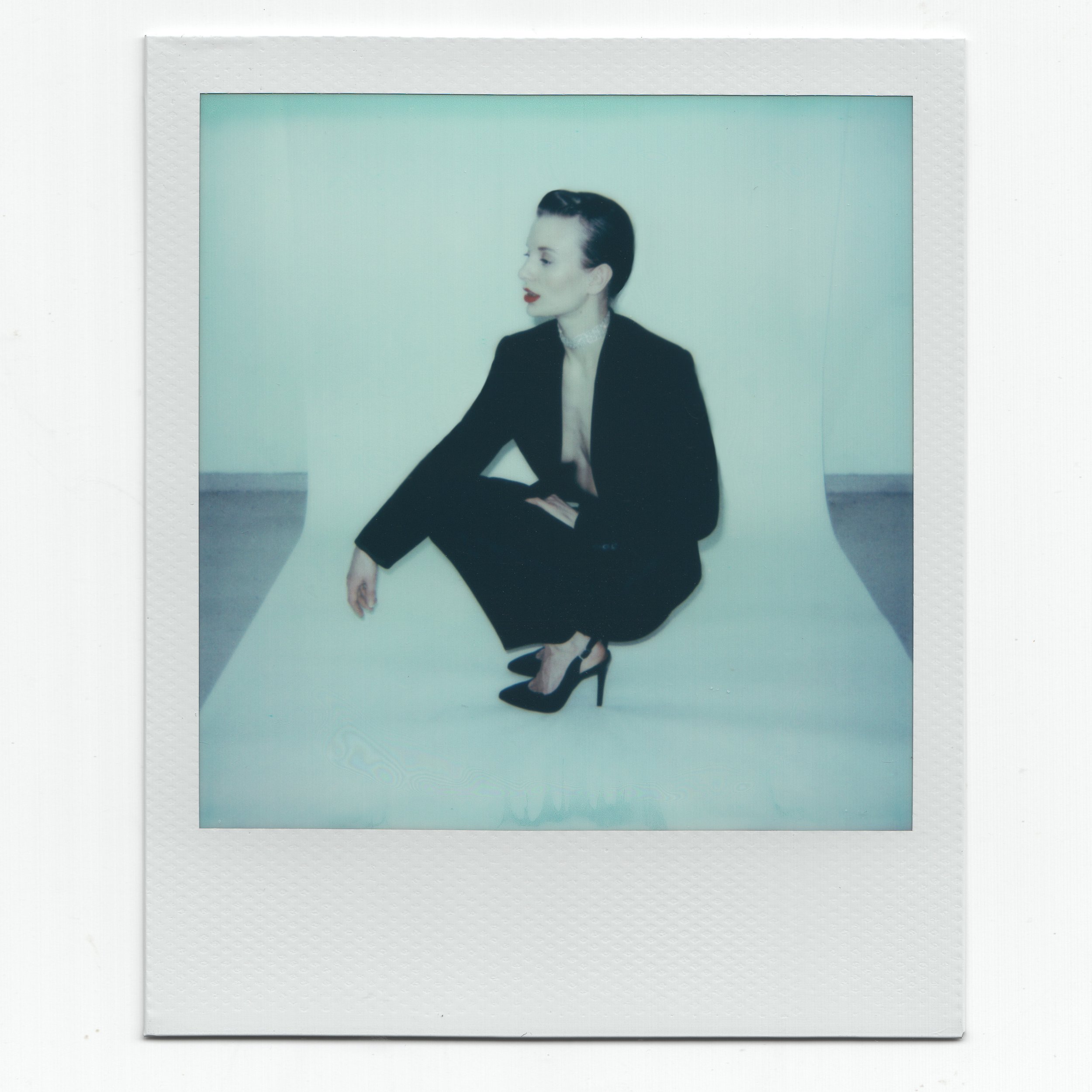 Model in a black suit and high heels squatting on a white backdrop, with a blurred vintage Polaroid effect.