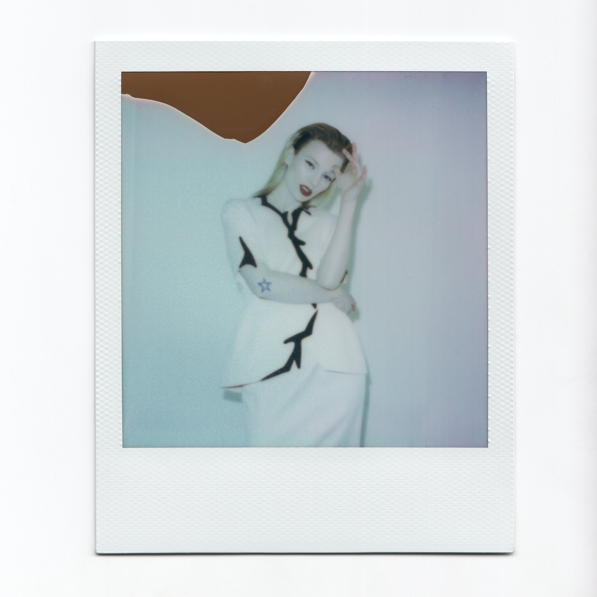 Polaroid photo of a model in a Thierry Mugler outfit with black zig-zag details, posing with a hand on head, against a plain background.