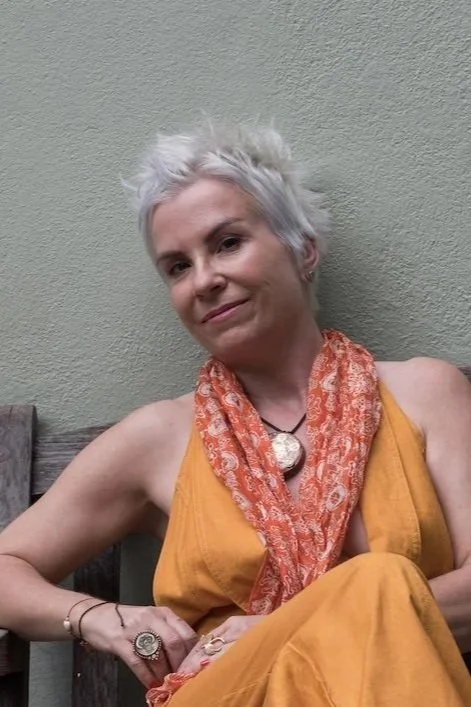 Woman with short white hair sitting against a green wall, wearing a yellow dress, orange scarf, and jewelry.