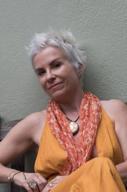 A woman with short, spiky white hair sitting against a plain wall, wearing a yellow top, an orange patterned scarf, and jewelry.