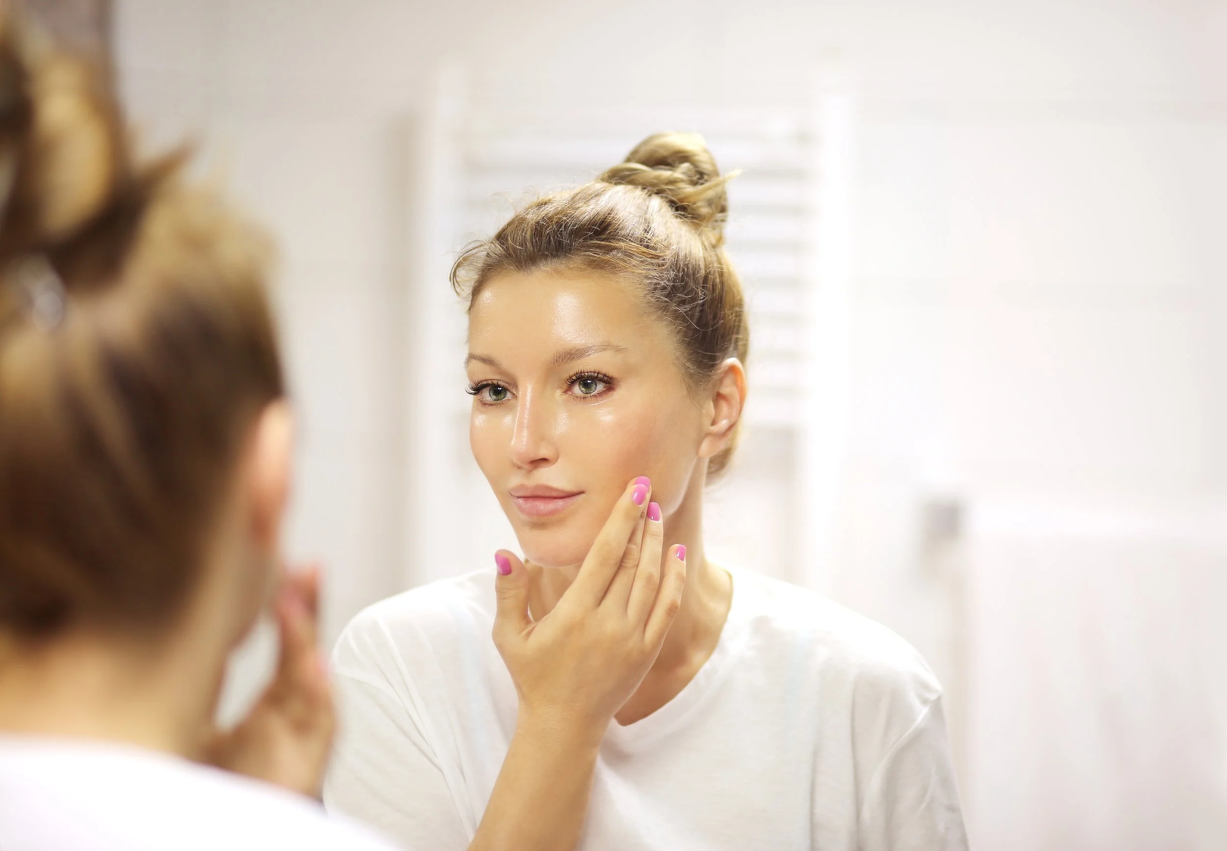 Woman in front of a bathroom mirror, touching her face gently, reflected in the mirror, with hair tied in a bun and wearing a white shirt.