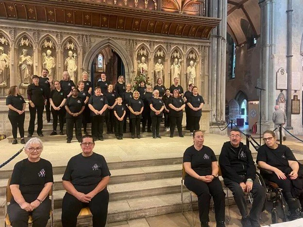 A choir group standing on stage inside a church, with raised hands and wearing black shirts, and four individuals seated in front of them.