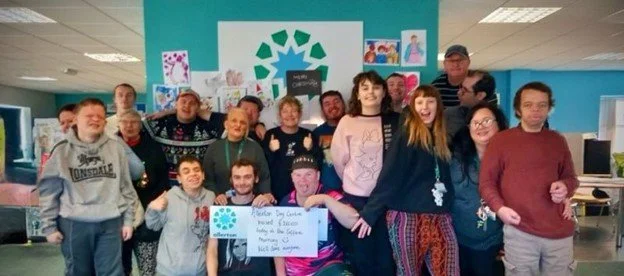 Group of people indoors, some holding a sign, smiling, with colorful artwork on the wall behind them.