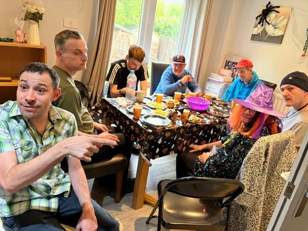 Group of people sitting around a dining table during a gathering, with food and drinks on the table, in a room with large windows and curtains, some wearing costumes or hats.