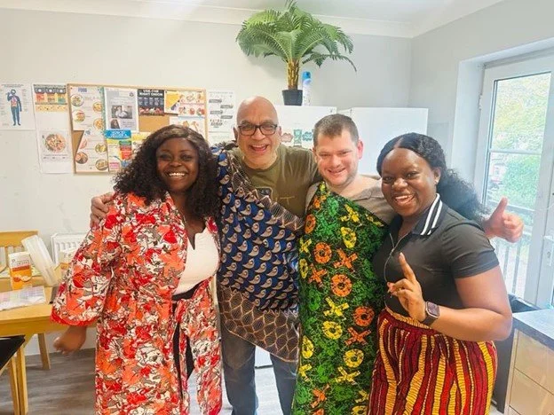 Group of four people smiling indoors, with one giving a thumbs-up, in a room with a window, posters on the wall, and a potted plant overhead.