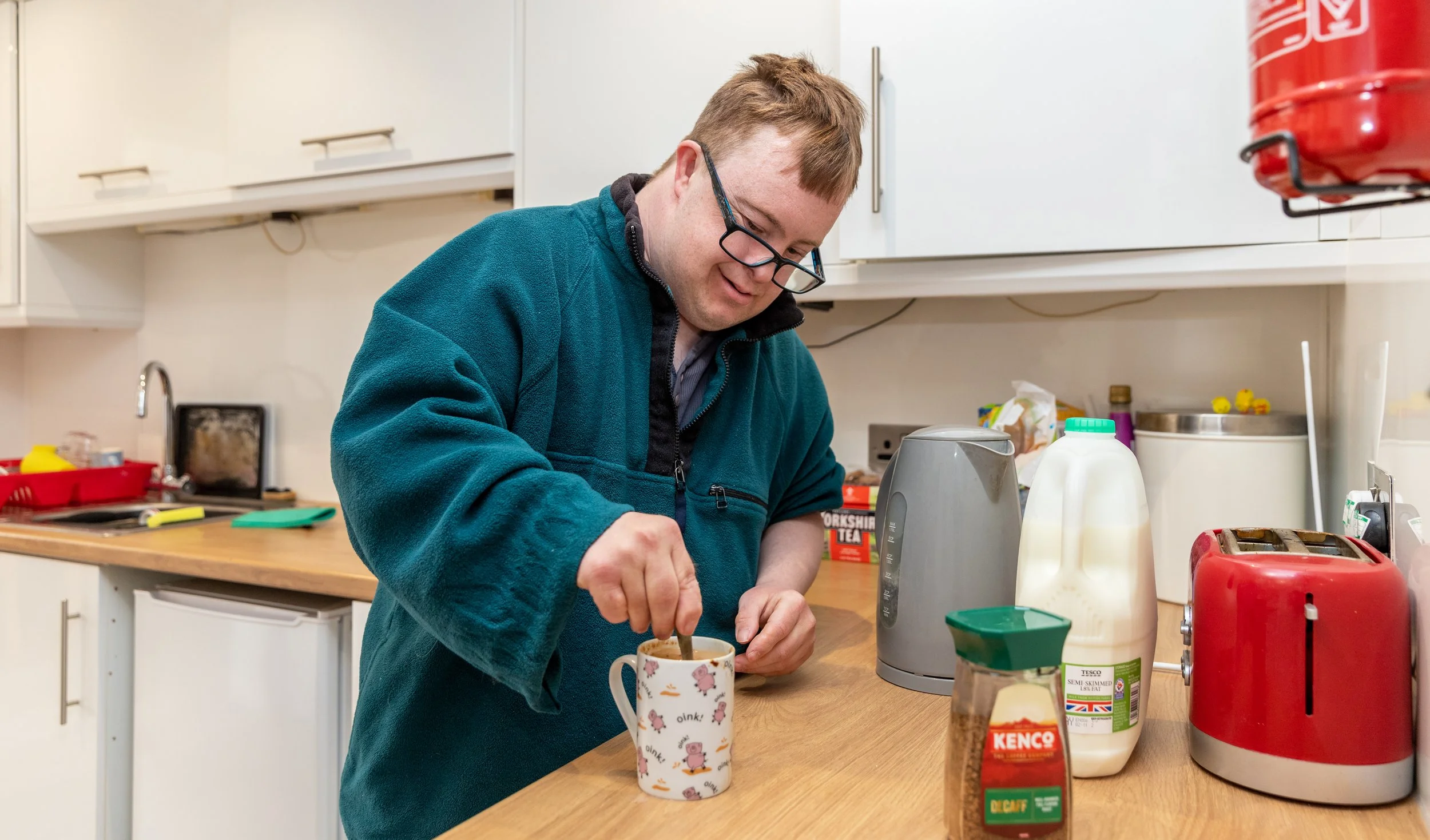 A man with glasses stirring a cup of tea or coffee in a kitchen.