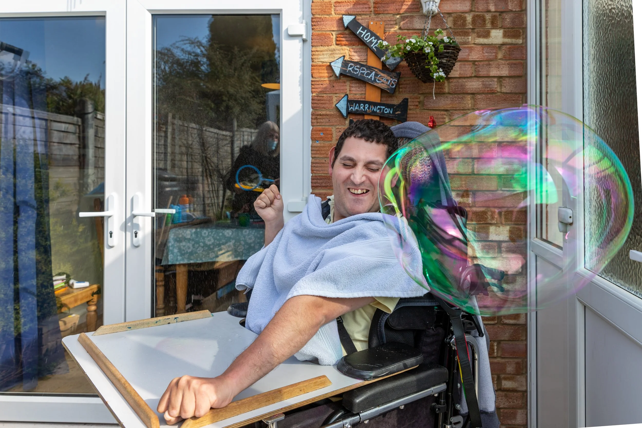 A man in a wheelchair is playing with soap bubbles outside a house, smiling, with a woman in the background through a glass door.