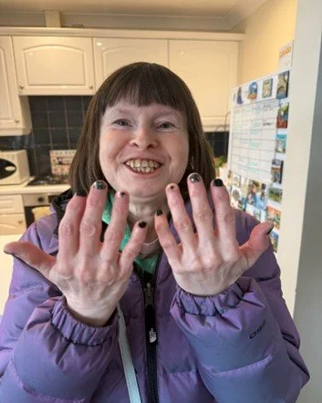 Person with short brown hair and braces showing their hands with black and green nail polish, smiling in a kitchen.