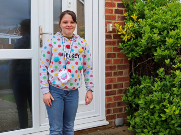 A young woman standing outdoors next to a brick wall and green bushes, wearing a gray hoodie with colorful polka dots and a lanyard with a small pouch, in front of a glass door.