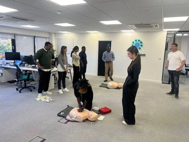 A group of people participating in a CPR training session in an office setting with computers and a logo on the wall.