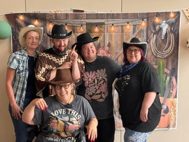 Five women posing in front of a western-themed backdrop with string lights, wearing cowboy hats and casual clothing.