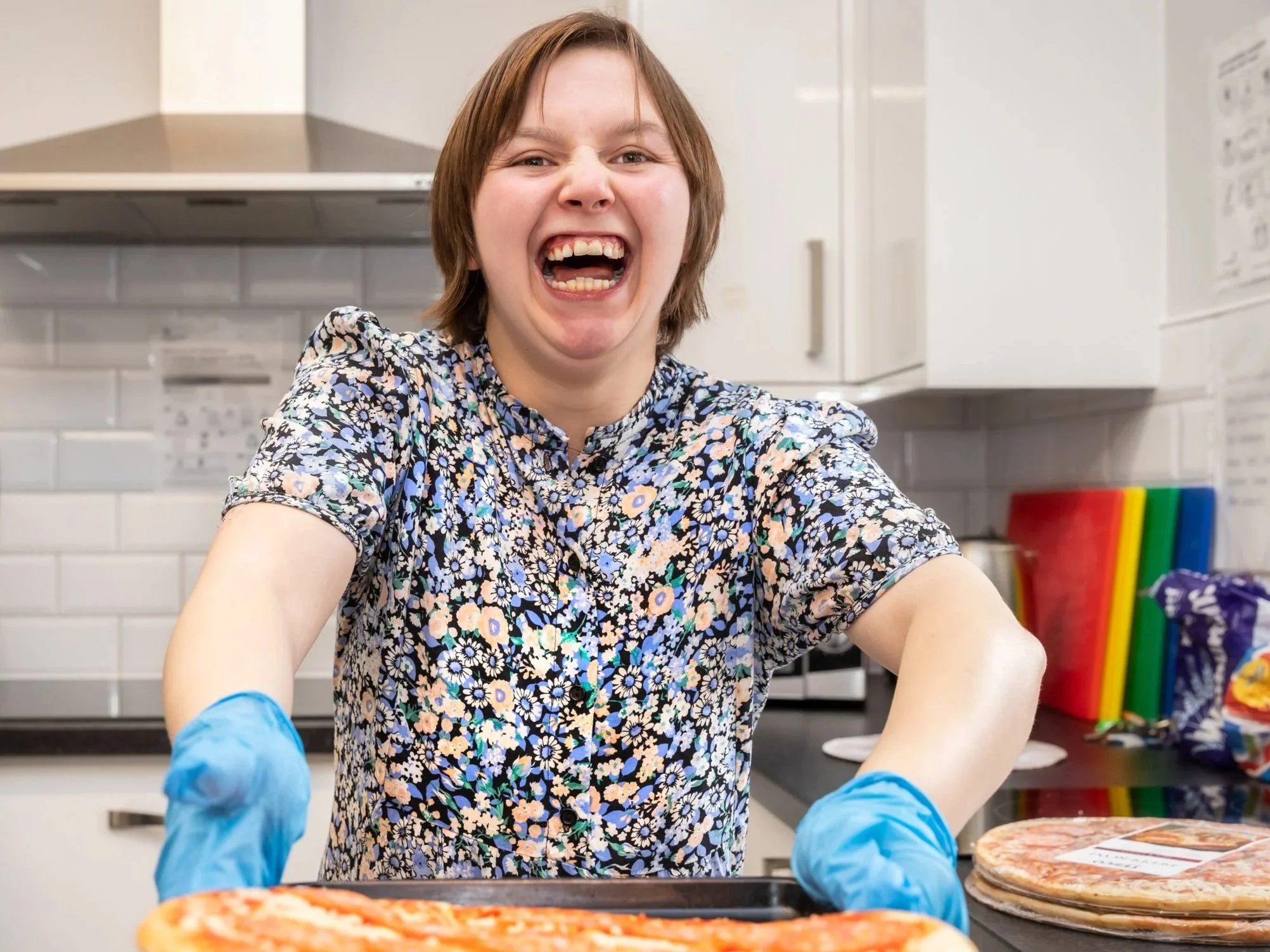 A woman with short brown hair and a floral shirt is laughing and wearing blue gloves. She is in a kitchen, standing over a tray of pizza, with kitchen cabinets, a microwave, colorful cutting boards, and snack bags in the background.