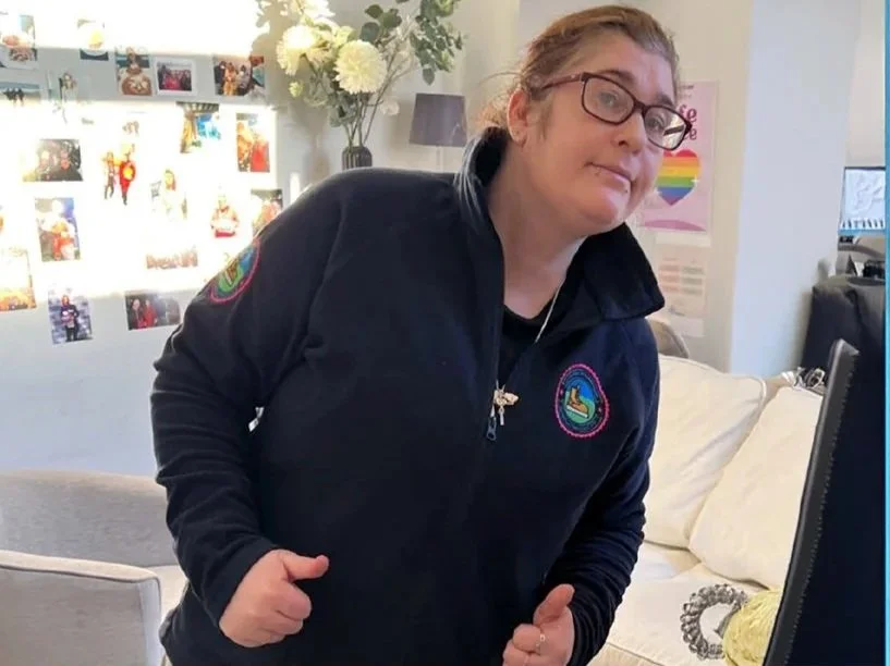 Woman with glasses, wearing a black jacket, leaning towards a computer, in a room decorated with photo collage, flowers, and posters.