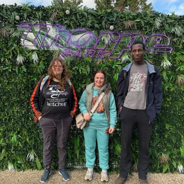 Three people posing in front of a lush green wall with a neon sign that says 'Hydeone Park'.