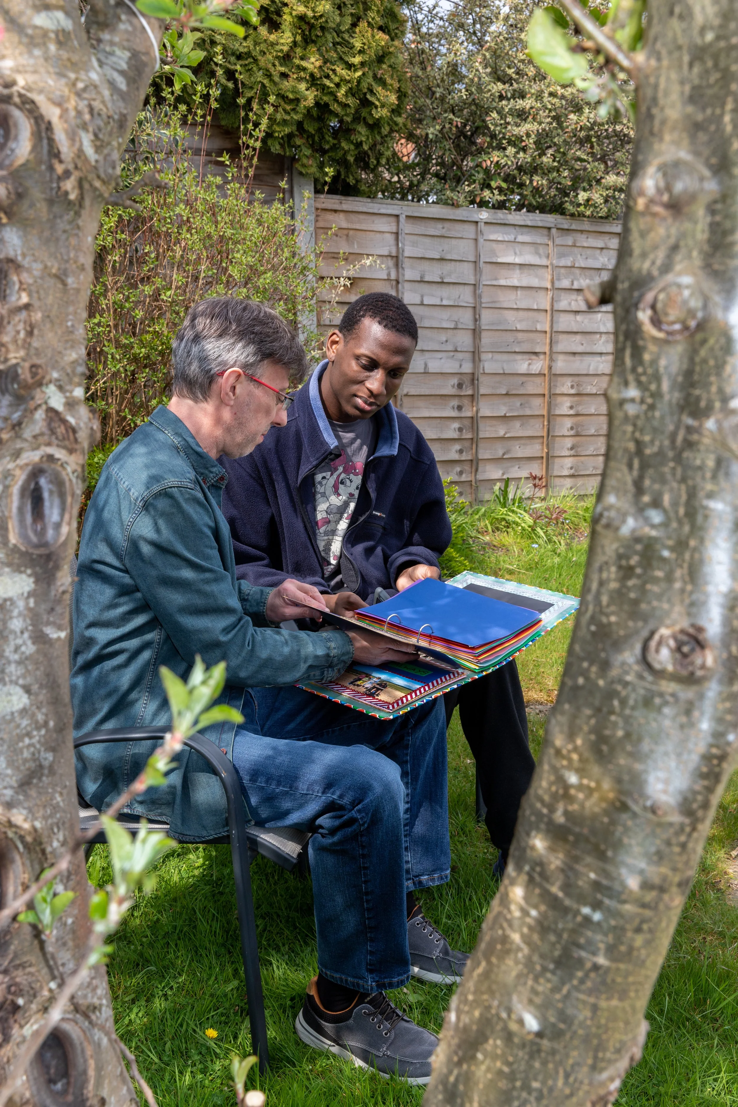 Two men sitting outdoors in a garden, looking at a photo album or scrapbook together. One is elderly with gray hair and glasses, and the other is a young adult with short black hair. They are surrounded by trees and bushes, with a wooden fence in the background.