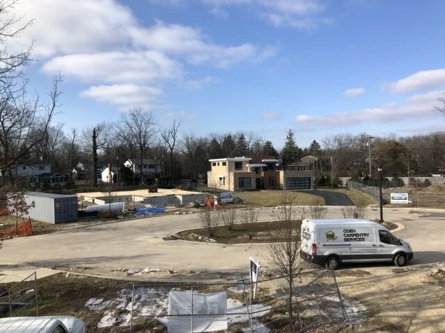 A residential construction site with a partially built house and a carpentry services van parked on a cul-de-sac under a partly cloudy sky.