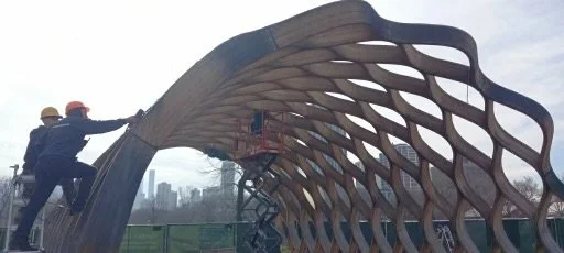 Two construction workers inspecting a wooden arch structure with lattice design at a park, with city skyline in the background.