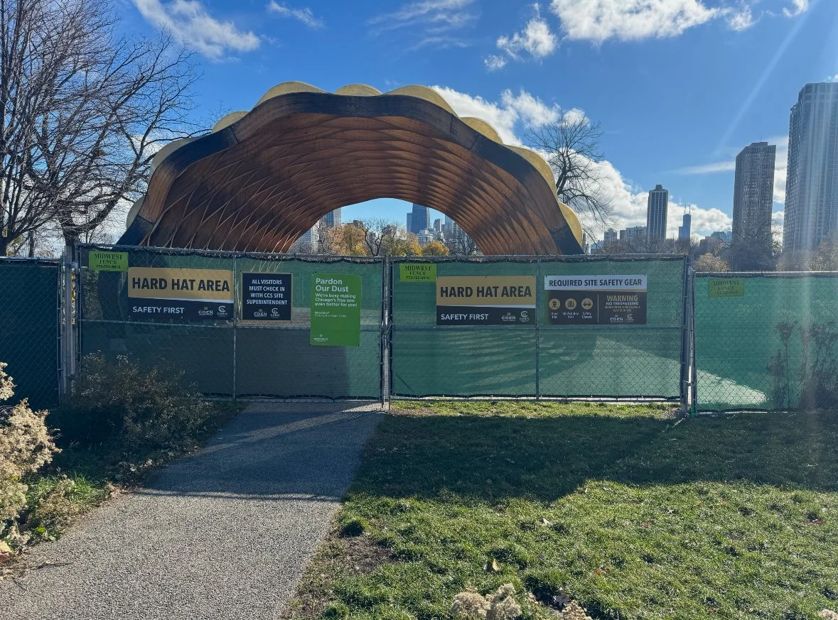 Construction site with a wooden arch structure, surrounded by green fencing and safety signage indicating a 'Hard Hat Area'. The background shows a clear sky and city skyline.
