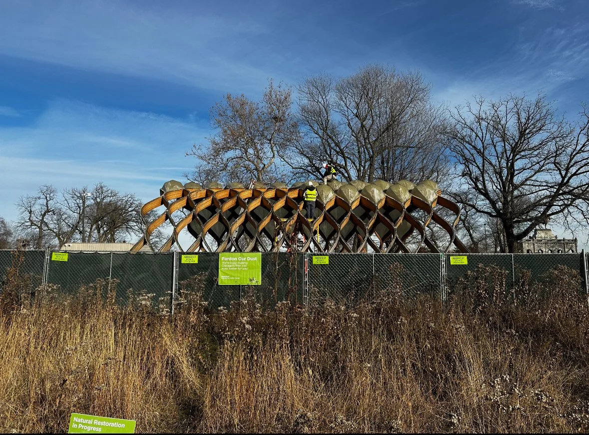 Construction workers on a geometric wooden structure behind a fence with signs reading 'Pardon Our Dust' and 'Natural Restoration in Progress'.