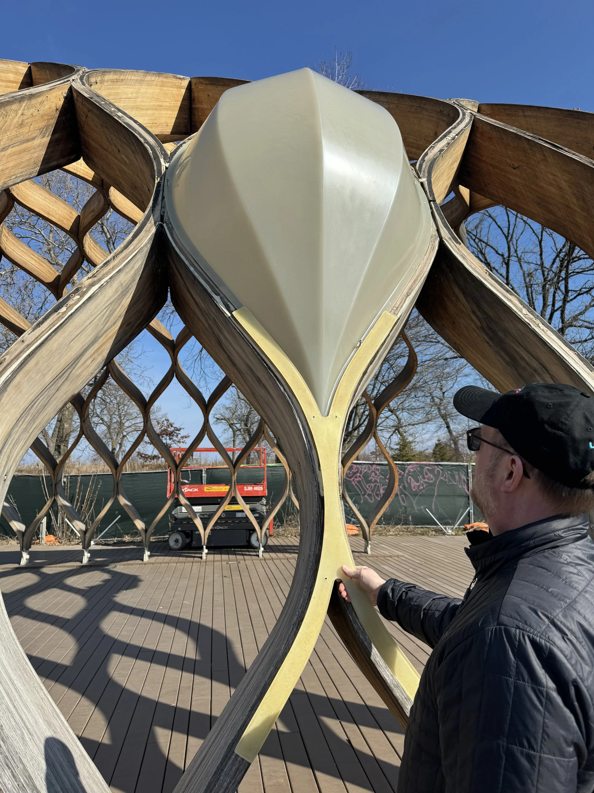 A person observes a large wooden and plastic geometric structure with a honeycomb pattern in a park. The sky is clear and blue.