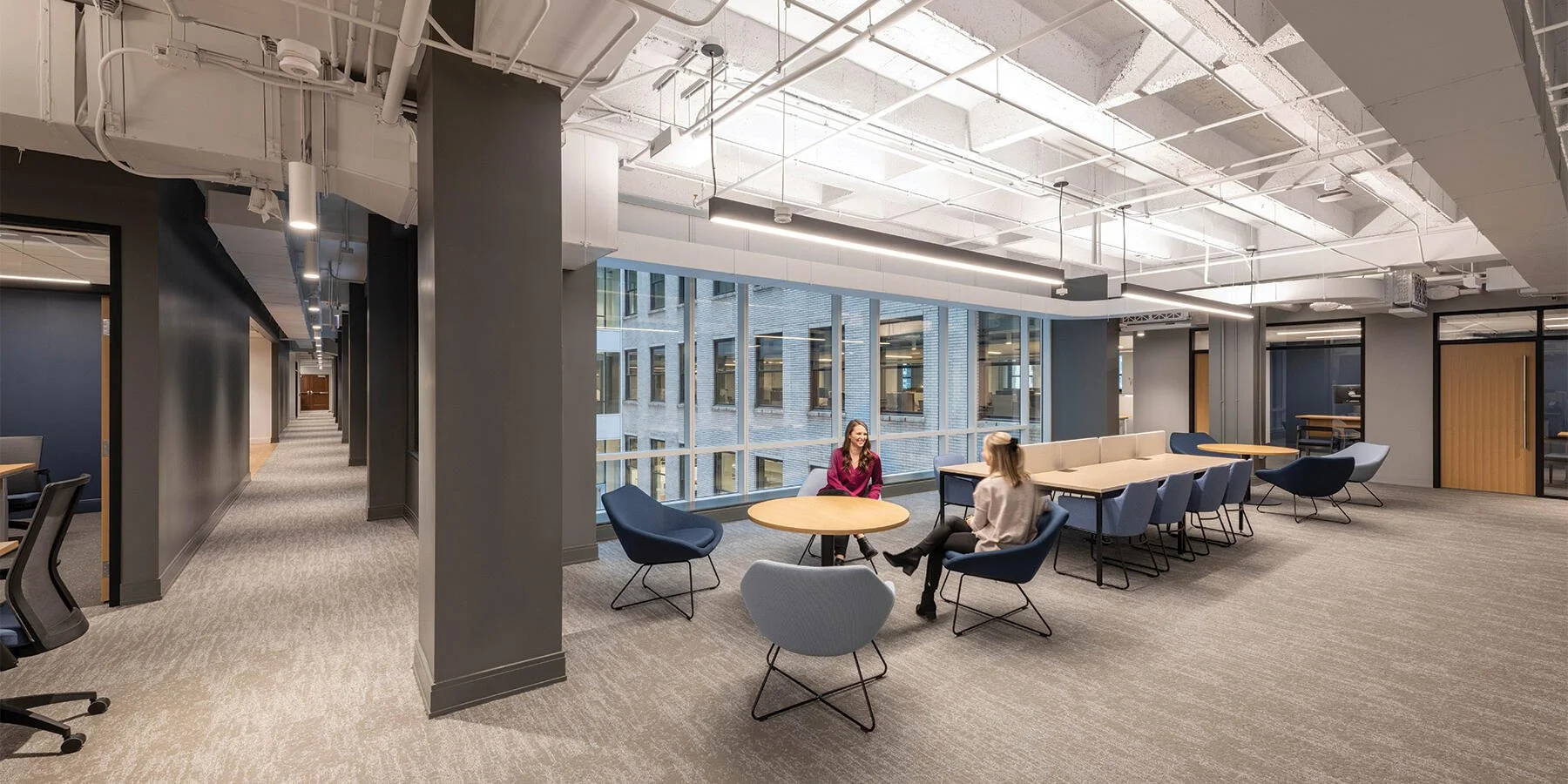 Modern office lounge area with large windows, round tables, and chairs, two women sitting and chatting.