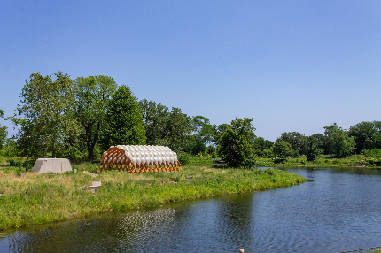 Scenic landscape with a pond, modern architectural pavilion, and trees under a clear blue sky.