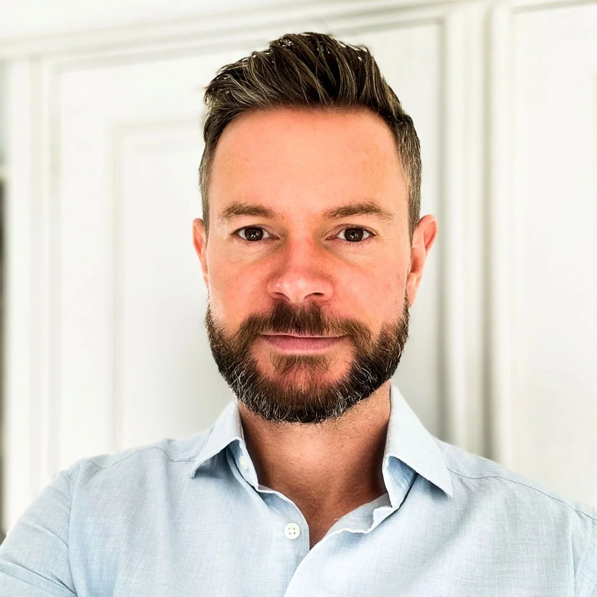 A man with dark hair and a beard, wearing a light blue collared shirt, standing indoors with a white door and trim in the background.