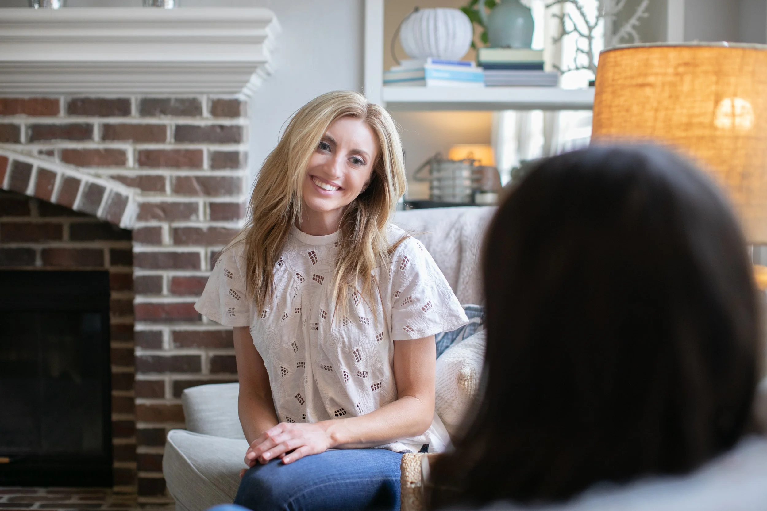 A smiling woman with long, wavy brown hair and blue eyes posing indoors in a living room with hanging plants, a large window, and a sofa with pillows in the background.