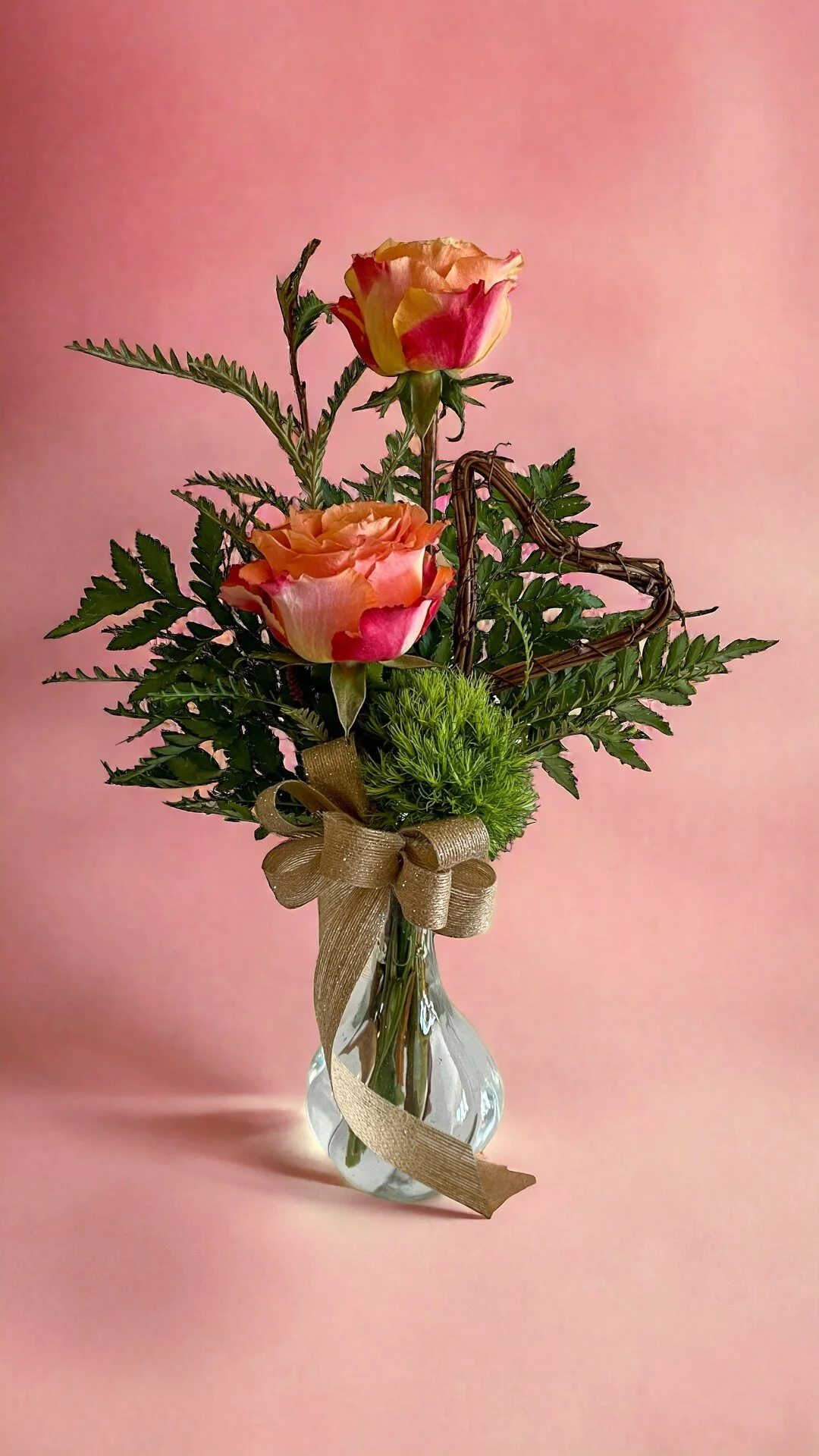 A small glass vase with a bouquet of pink and peach roses, green ferns, and decorative greenery, tied with a gold ribbon on a pink background.