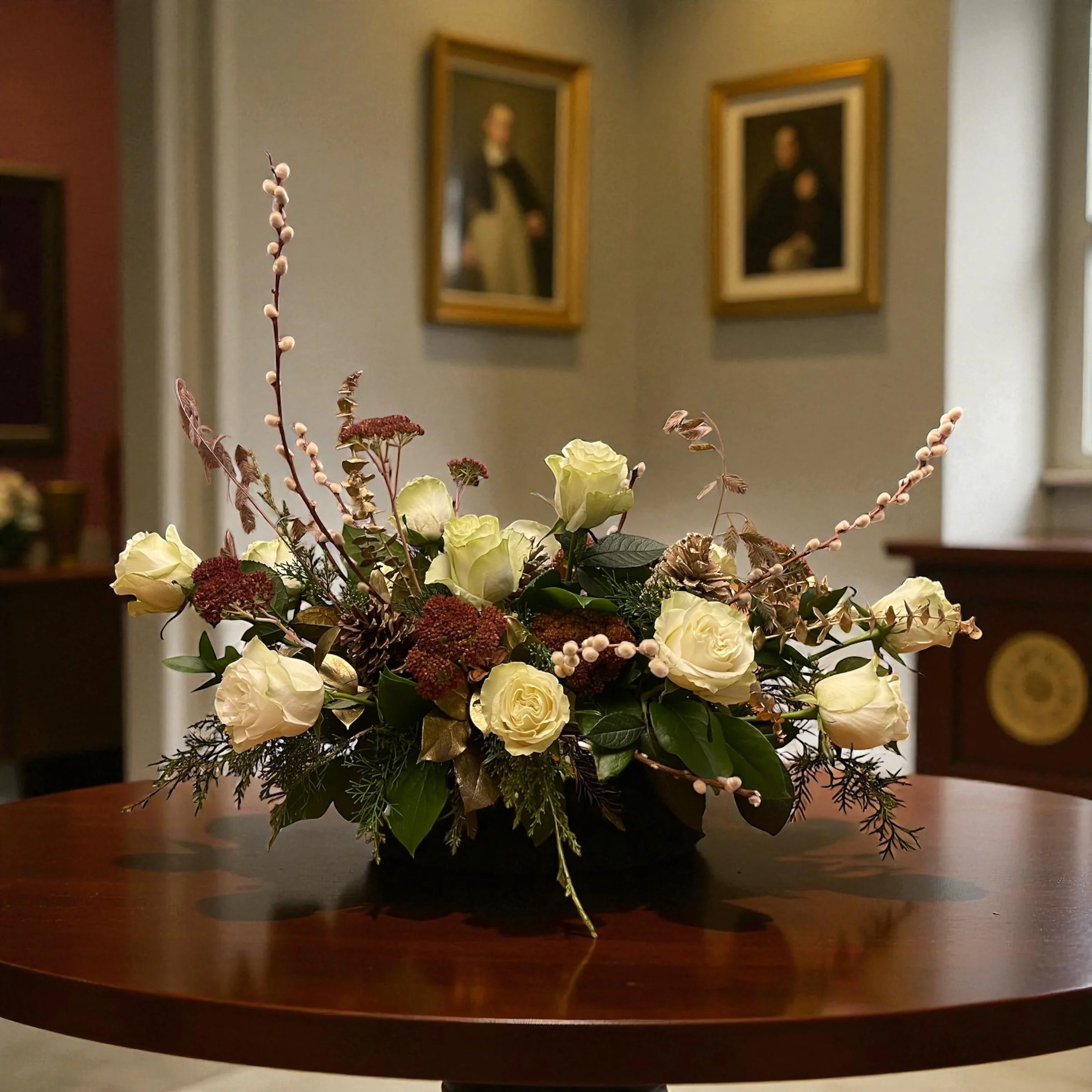 A floral arrangement with white roses, red filler flower, and greenery on a wooden table in a room with framed portraits on the wall.