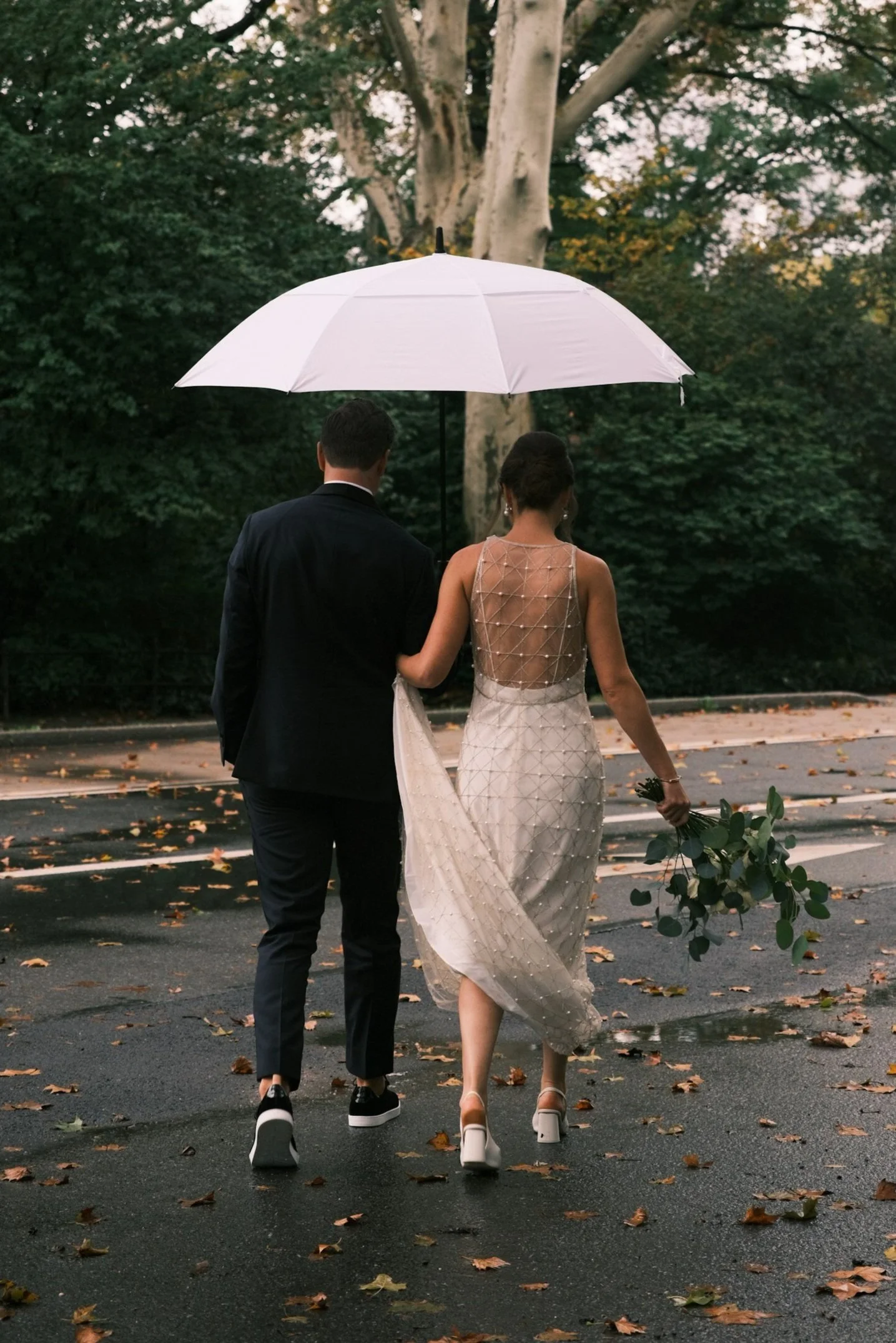 A couple dressed in wedding attire walking together under a white umbrella on a rainy day, with fallen leaves on the road and trees in the background.