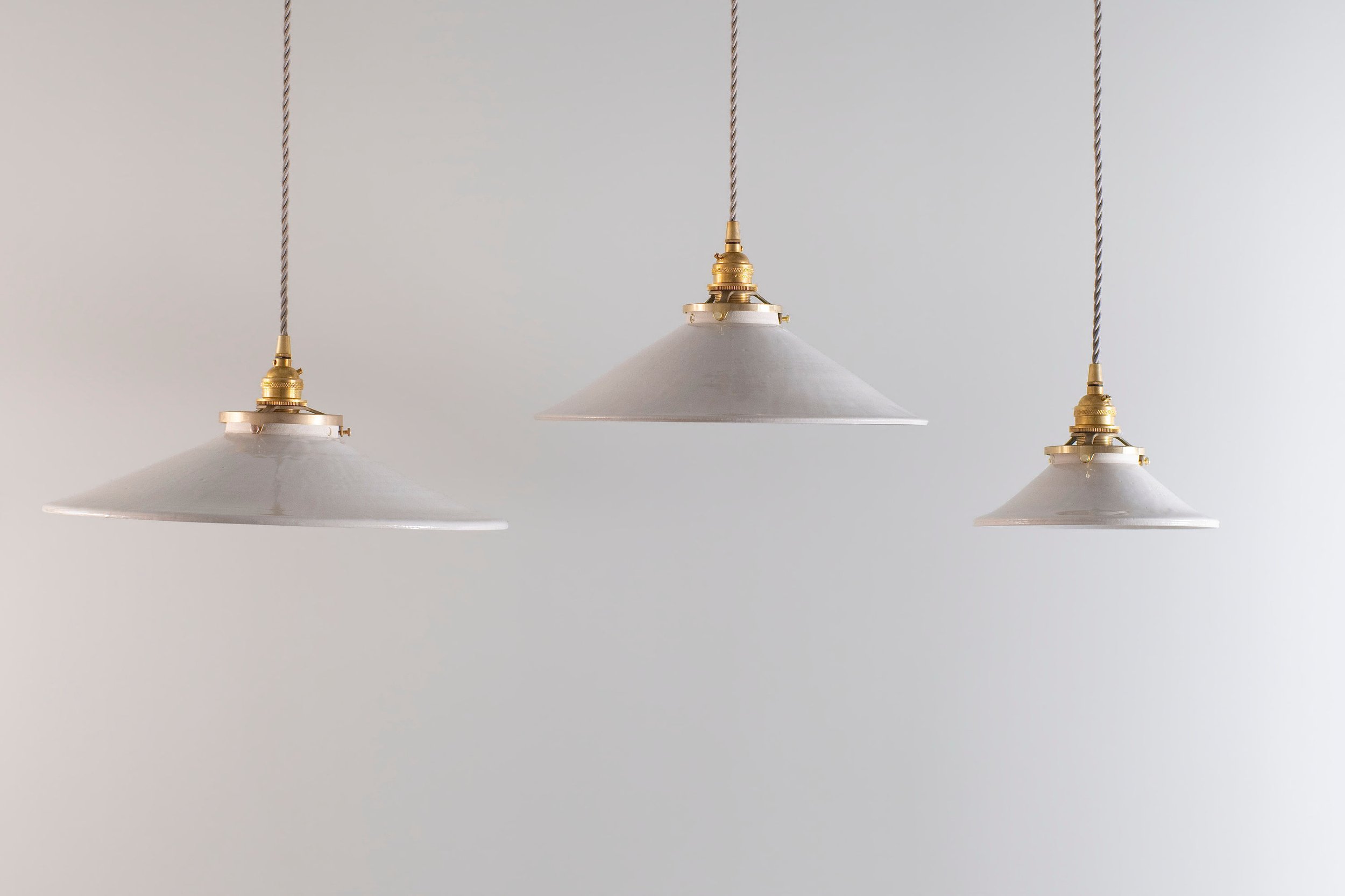 Three sizes of white, ceramic, handmade pendant lights with brass accents hanging from ceiling on antique braid cords.