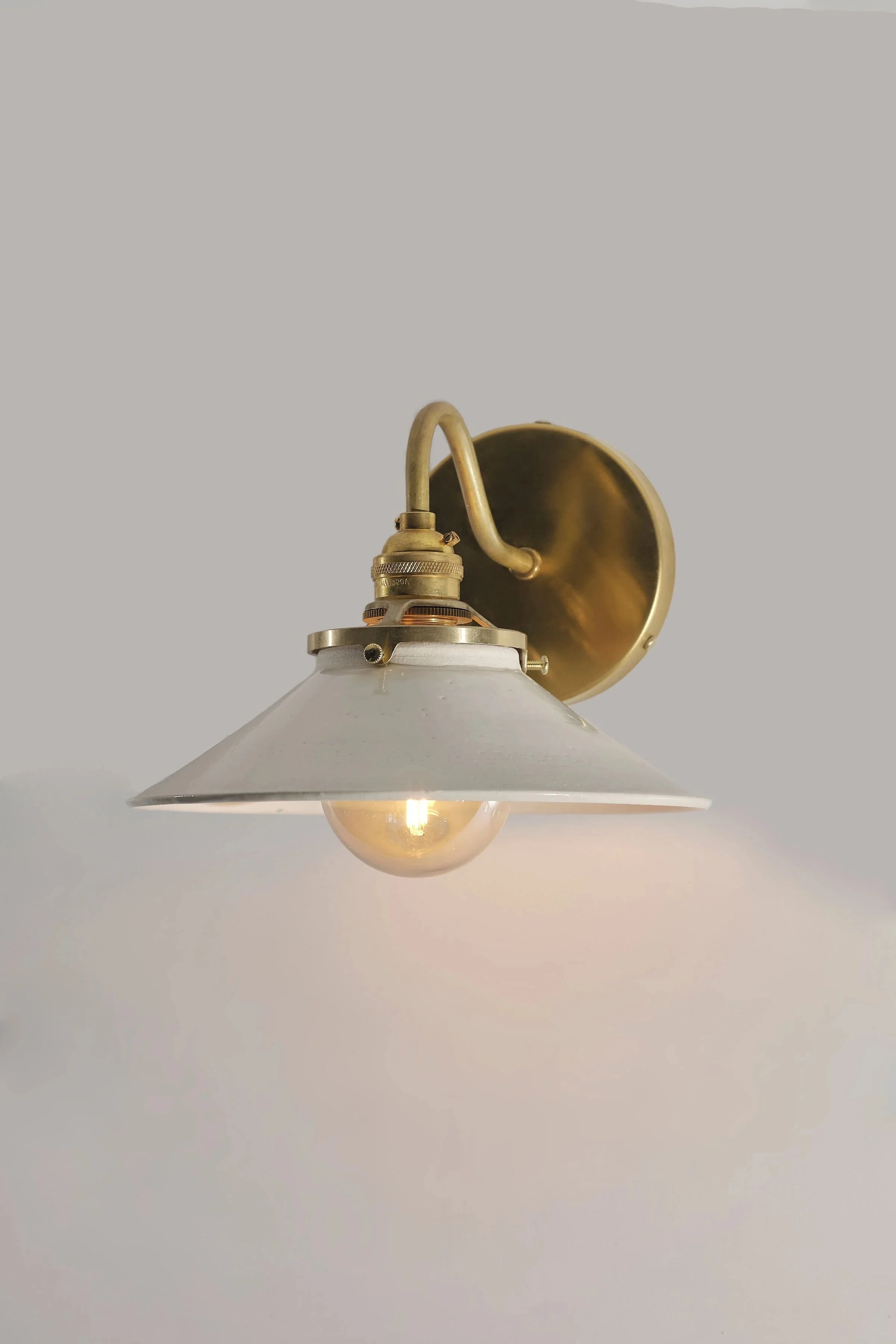 A vintage brass wall-mounted lamp with a white handmade ceramic lampshade and an exposed light bulb, against a plain light background.
