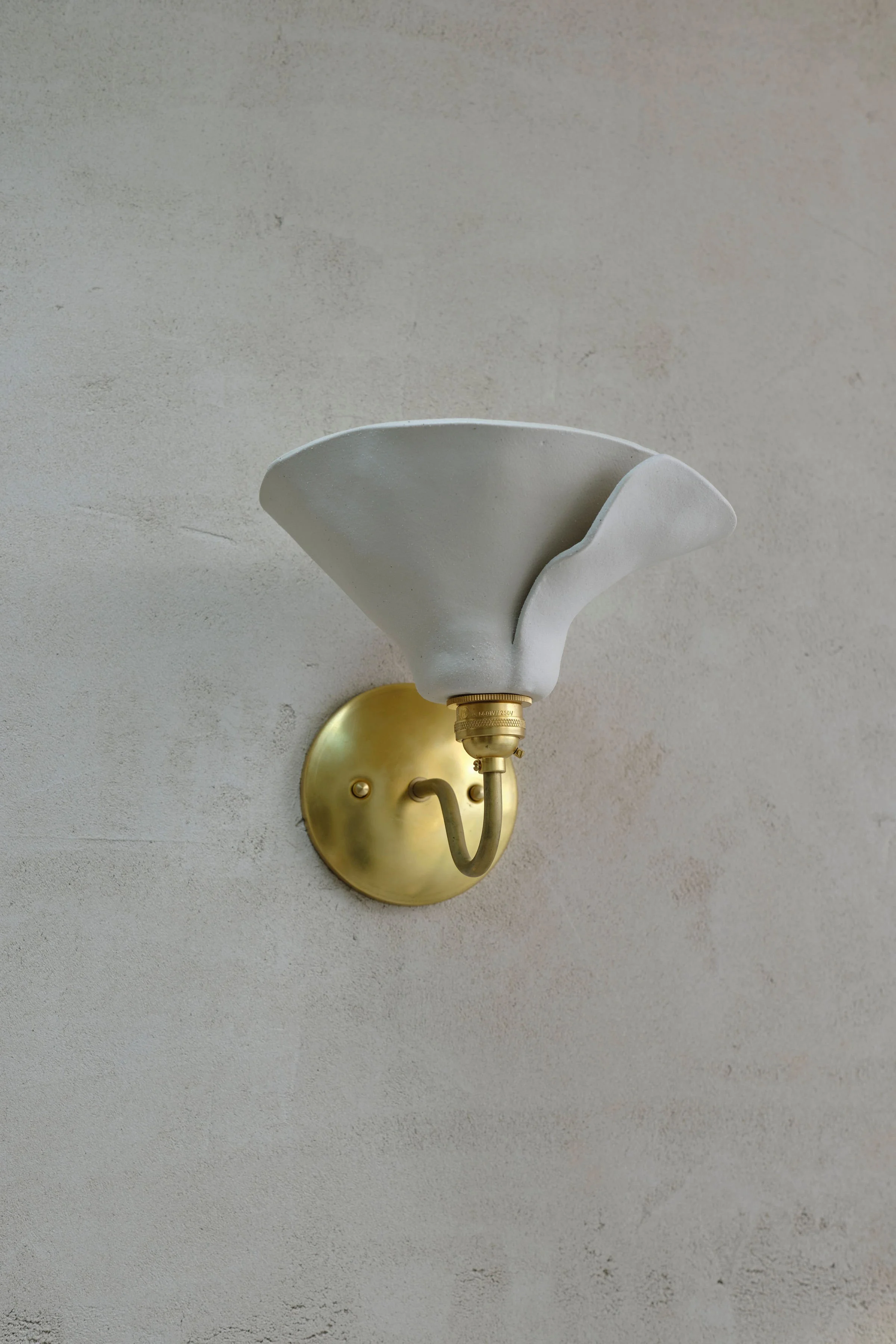 Wall-mounted light fixture with a gold base and a white, flower-shaped shade against a textured beige wall.