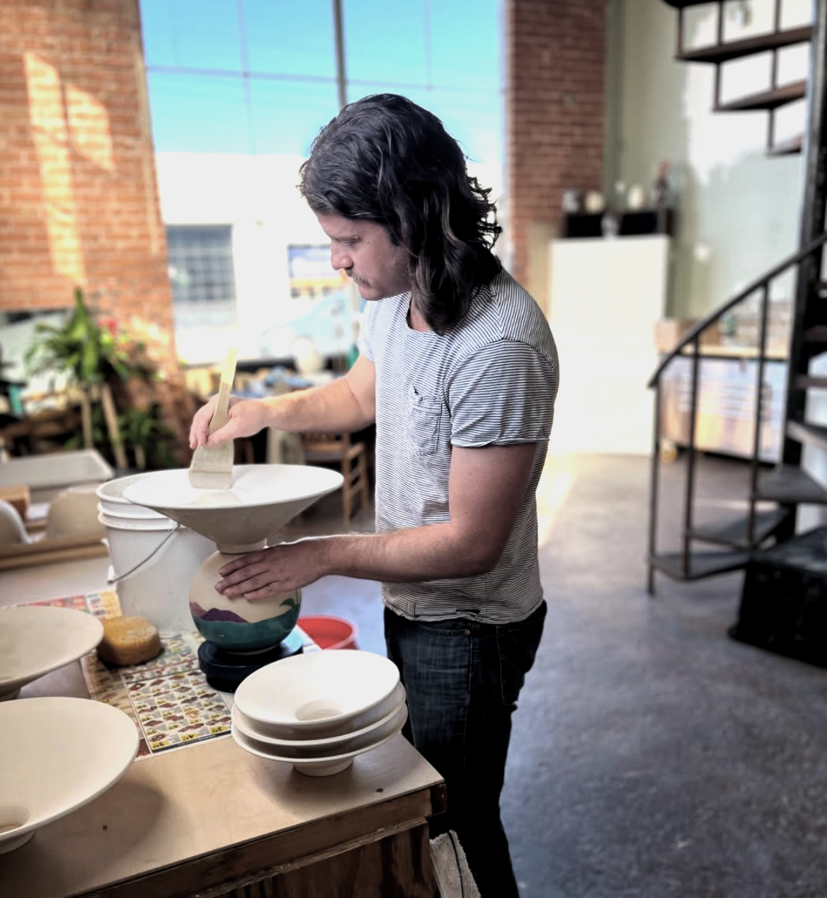 Ceramic artist with long hair in a striped shirt glazing handmade ceramic light l in a bright, industrial-style studio with brick walls and stairs.