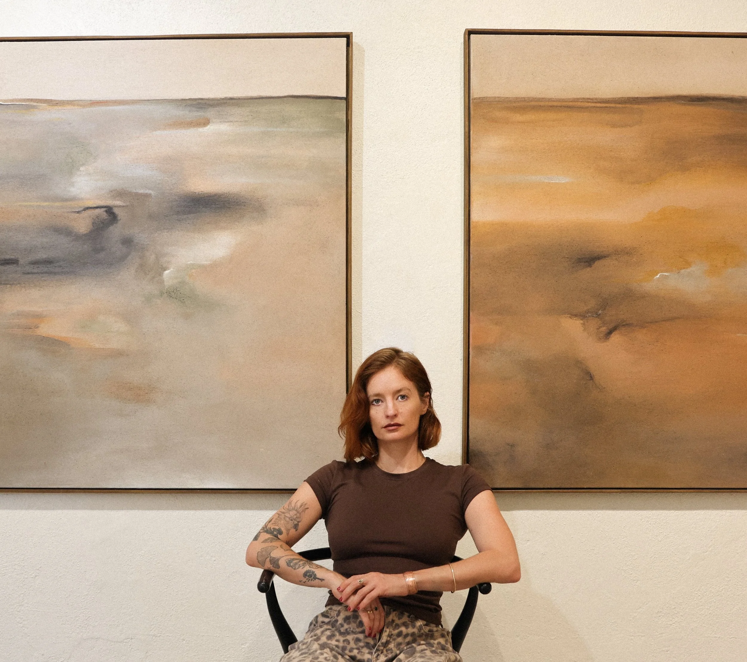 A woman with reddish-brown hair and tattoos on her arms sitting in front of two abstract landscape paintings on a beige wall, wearing a brown t-shirt and camouflage pants.