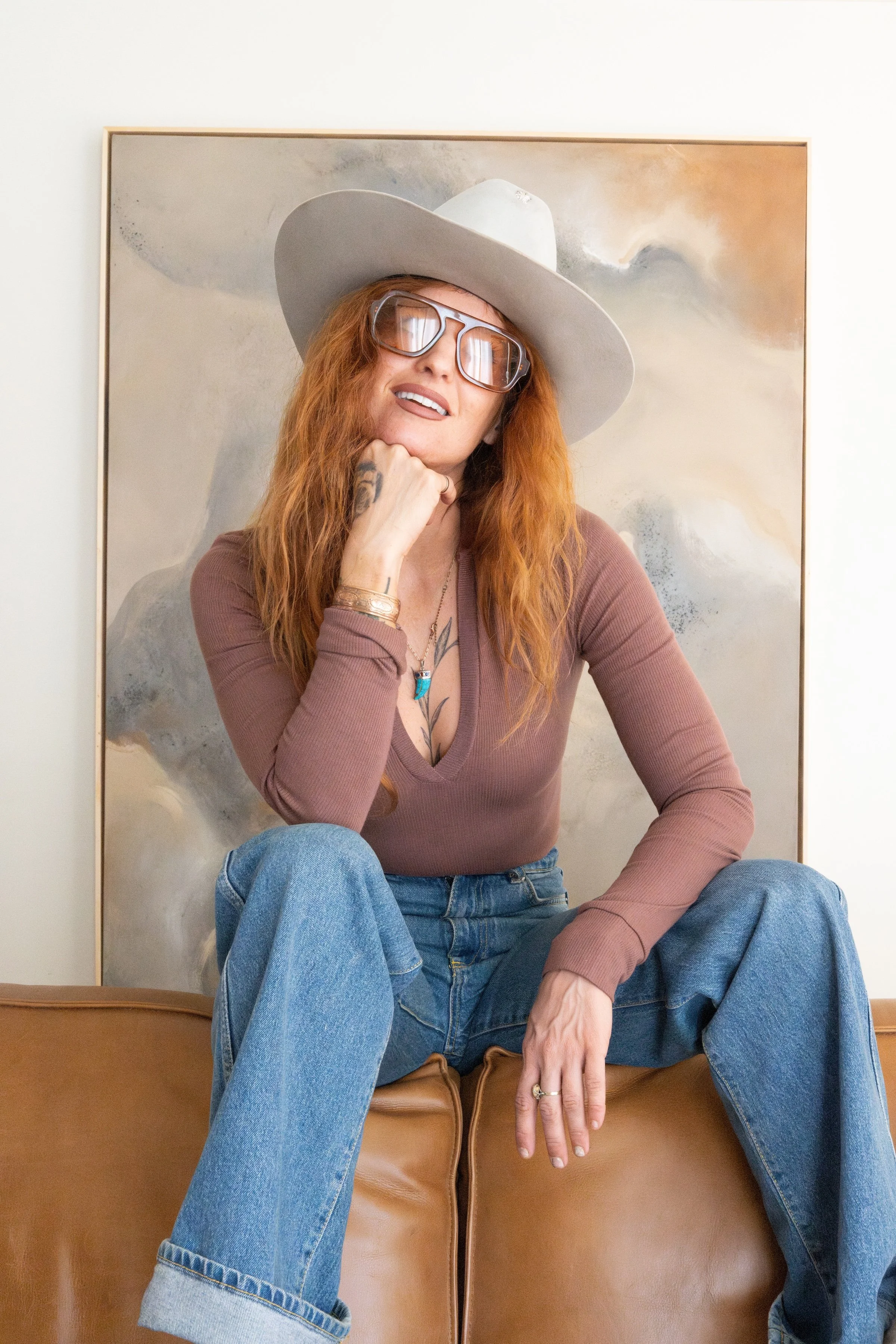 Woman with long red hair, wearing a gray wide-brimmed hat and large glasses, sitting on a leather chair against an abstract painting background. She is dressed in a mauve long-sleeved top, blue jeans, and has a tattoo on her chest, jewelry on her left wrist, and rings on her fingers. She is smiling and posing with her hand resting on her chin.
