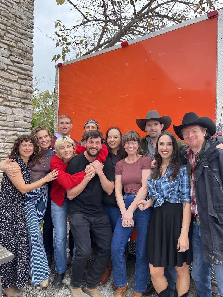 Caroline Rose with the cast and crew in western wear and cowboy hats in front of an orange trailer on location