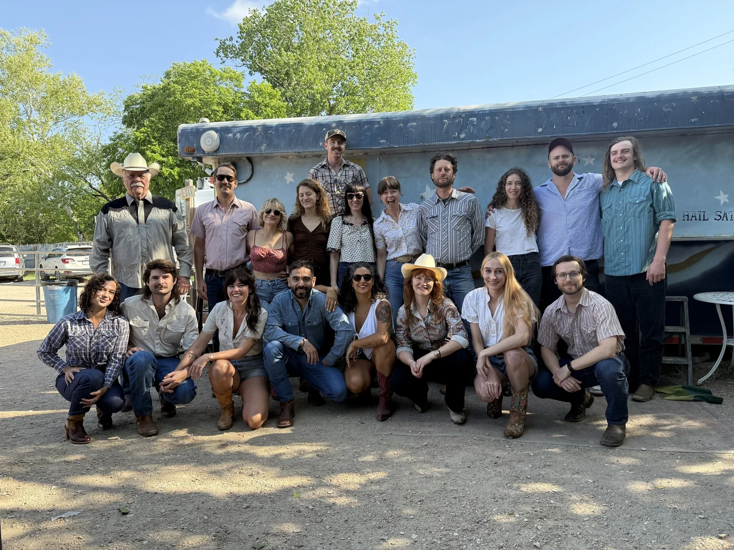 Nineteen country dancers in western shirts, boots, and cowboy hats posed together in front of a weathered blue trailer on the location of the Spec's commercial shoot